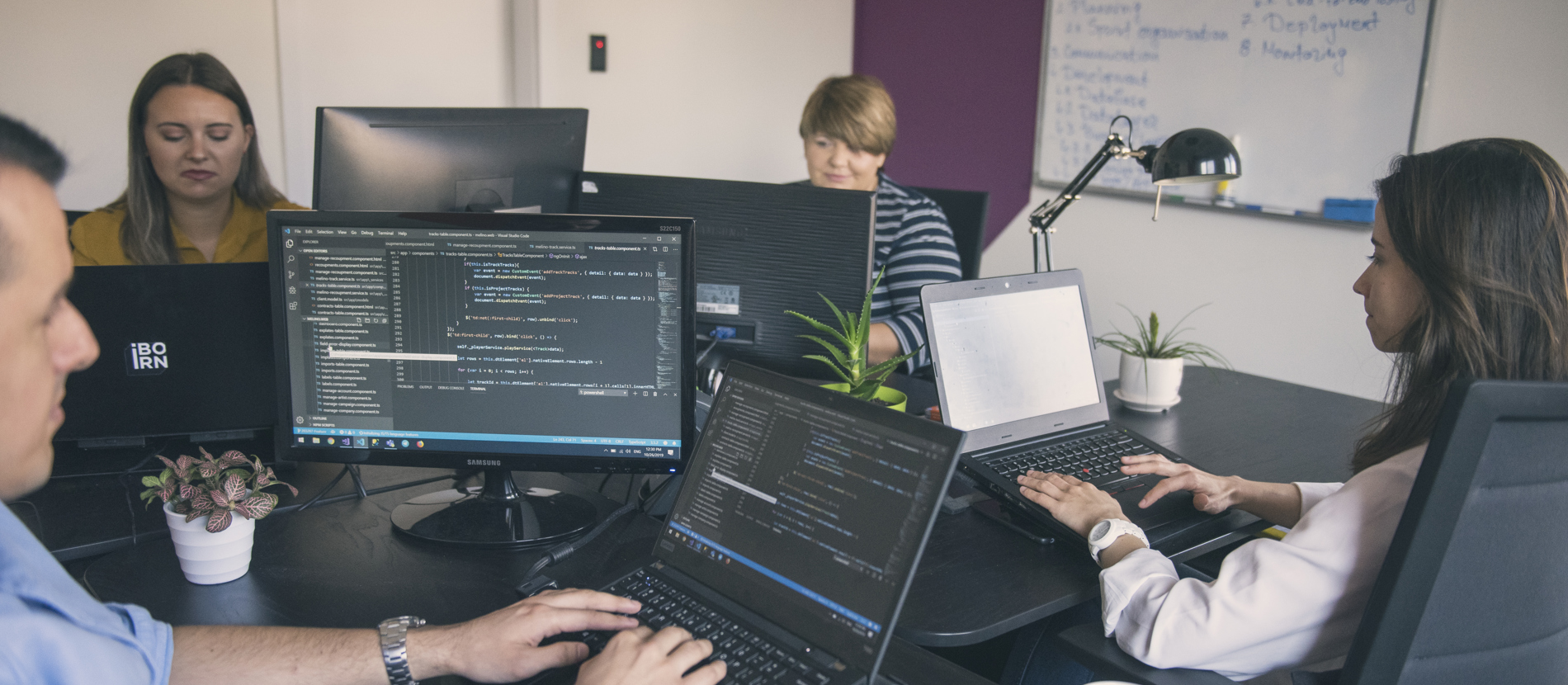 Close-up of computer and laptop screens with people working in the background.