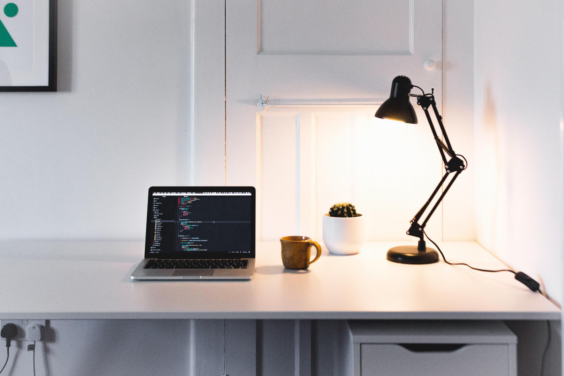 Laptop on a desk with a coffee mug, a small cactus, and a desk lamp.  