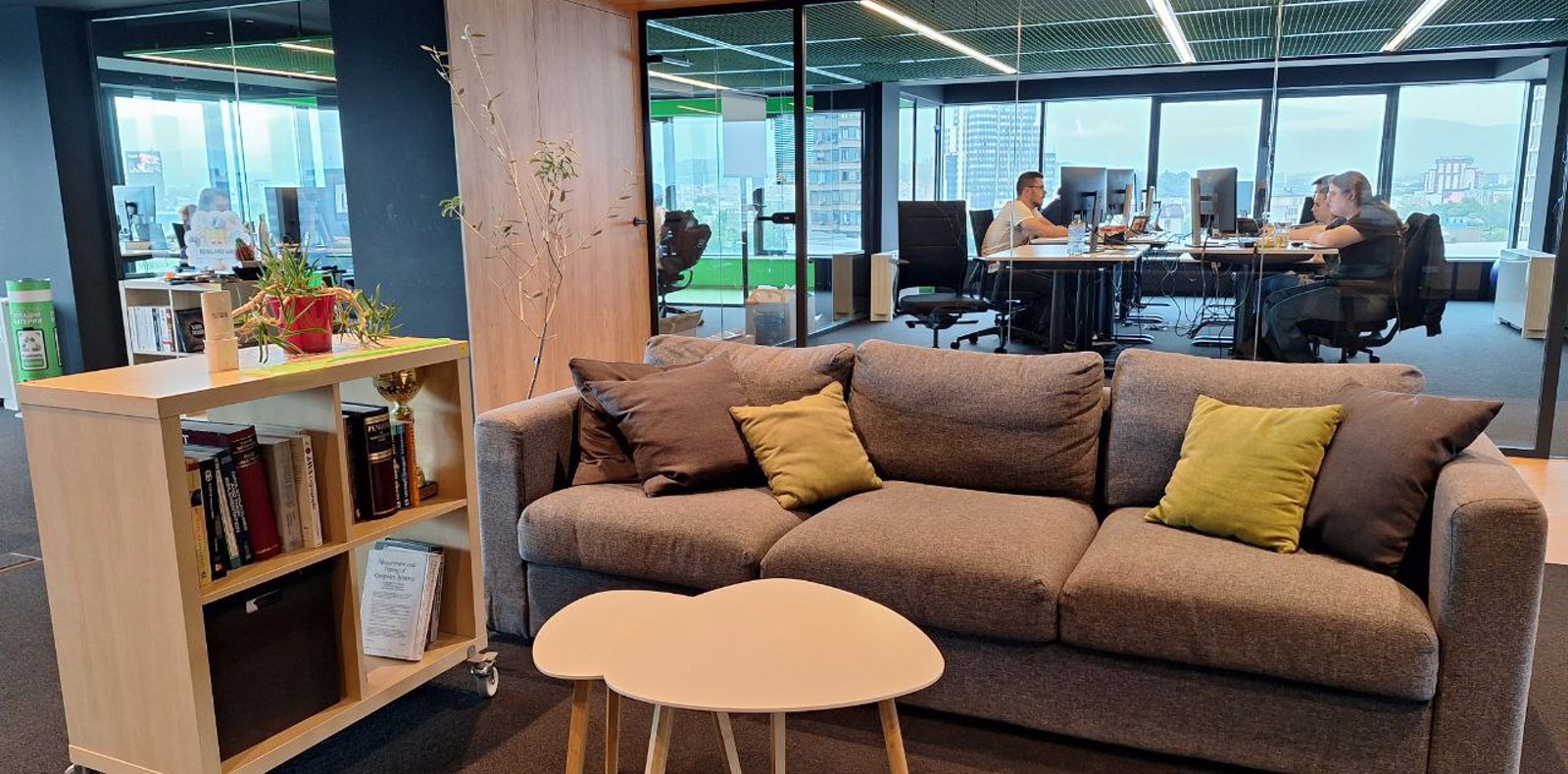 Image of a contemporary office featuring a couch, table, and chairs, and people working in the background offices.