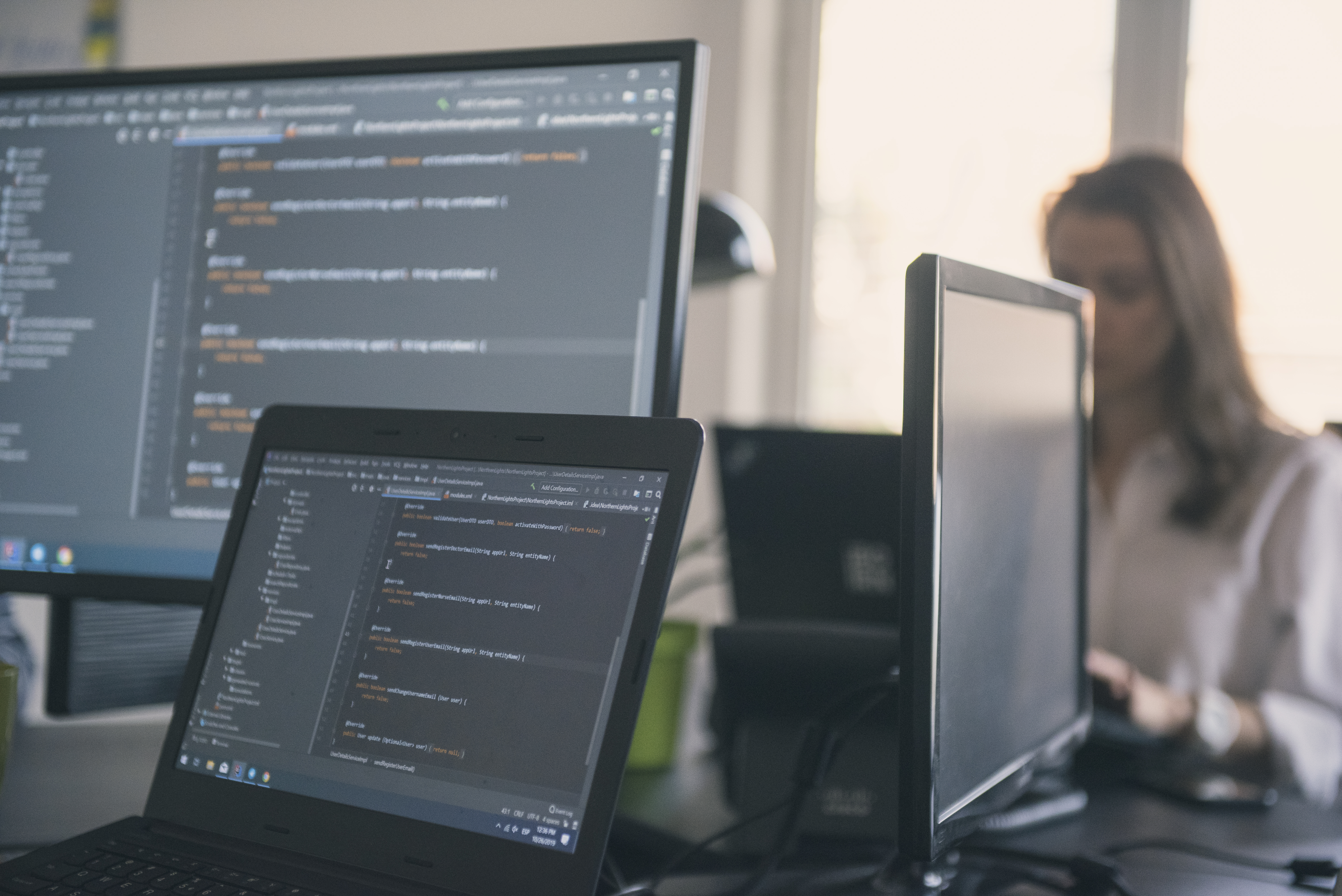 Close-up of computer and laptop screens with people working in the background.