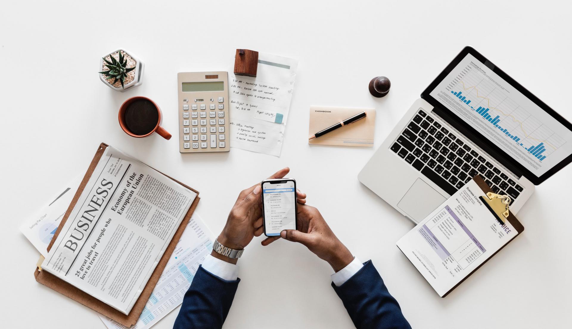 A person holding a mobile phone with a laptop, calculator, newspaper, and cup of coffee and succulent in front of him. 